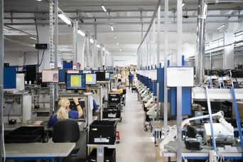 Moderne Produktionshalle mit Mitarbeitern an Arbeitsplätzen und Computern, die elektronische Komponenten montieren.