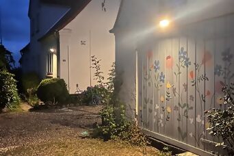 Ein sanft beleuchteter Weg neben einem Haus mit einer bunten Blumenwand an einem Garagentor, umgeben von Grün unter einem dämmrigen Himmel.