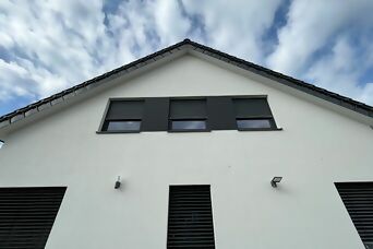 Moderne Hausfassade mit Steinel Außenleuchten und dunklen Fensterläden unter blauem Himmel.